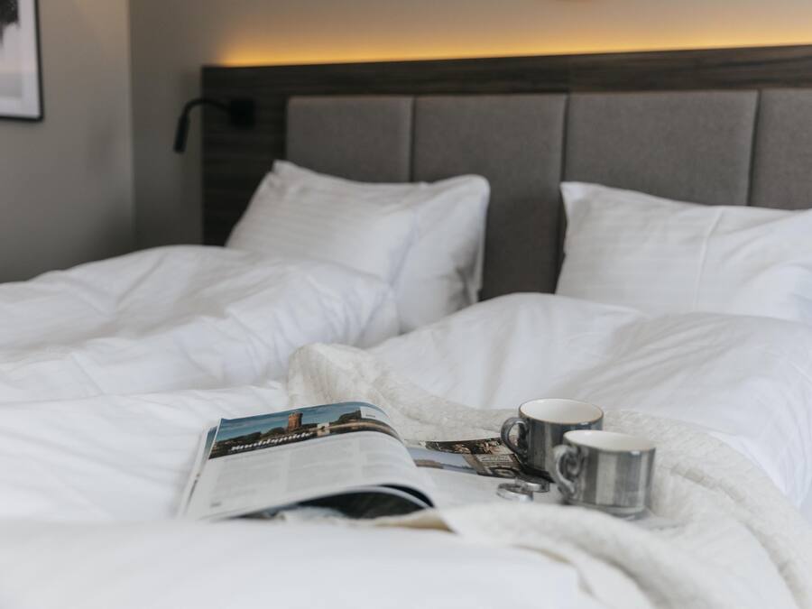 Una cama bien tendida con sábanas blancas, una revista y una taza sobre la cama.