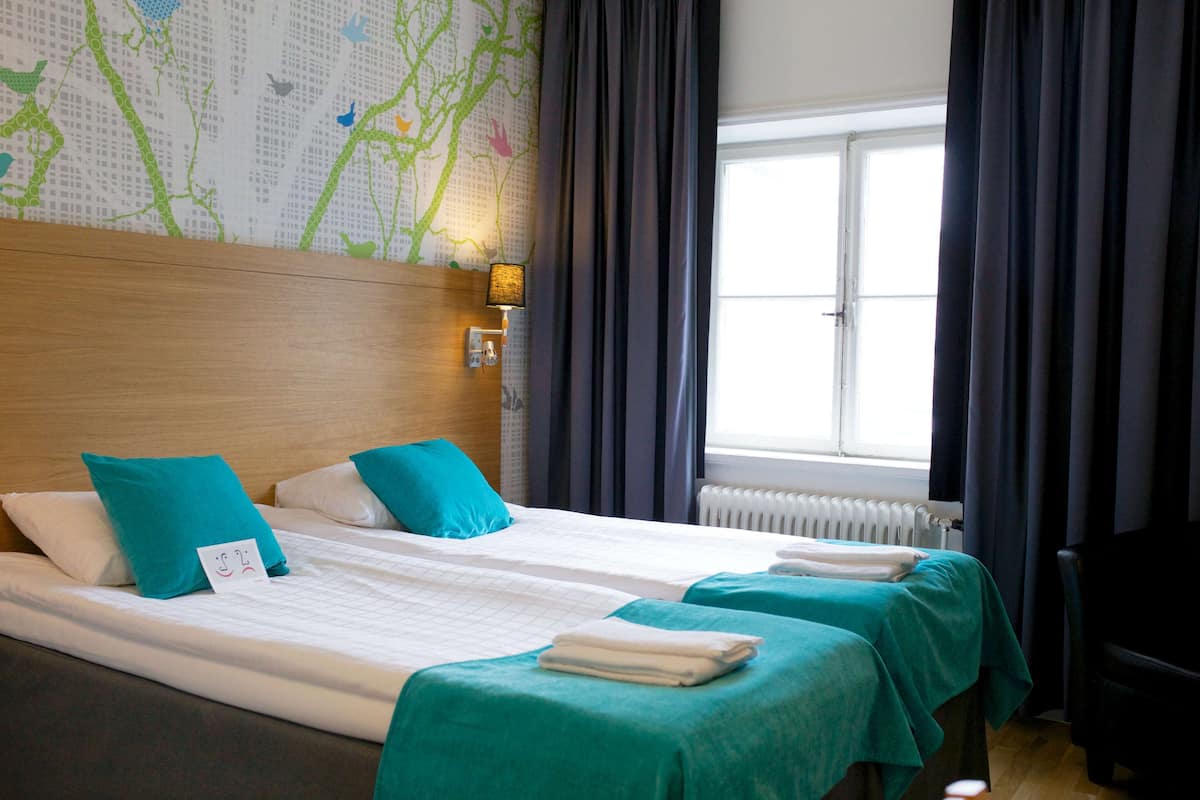 A hotel room with two beds, teal bedspreads, a wooden headboard, a map wall, a window with curtains, and a radiator.