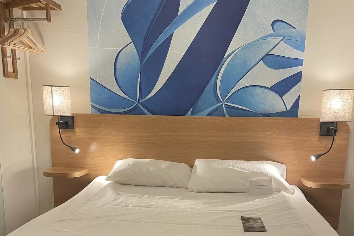 A hotel room with a wooden headboard, two bedside lamps, a large abstract painting on the wall, a neatly made bed with white linens, and a folded towel on the bed.