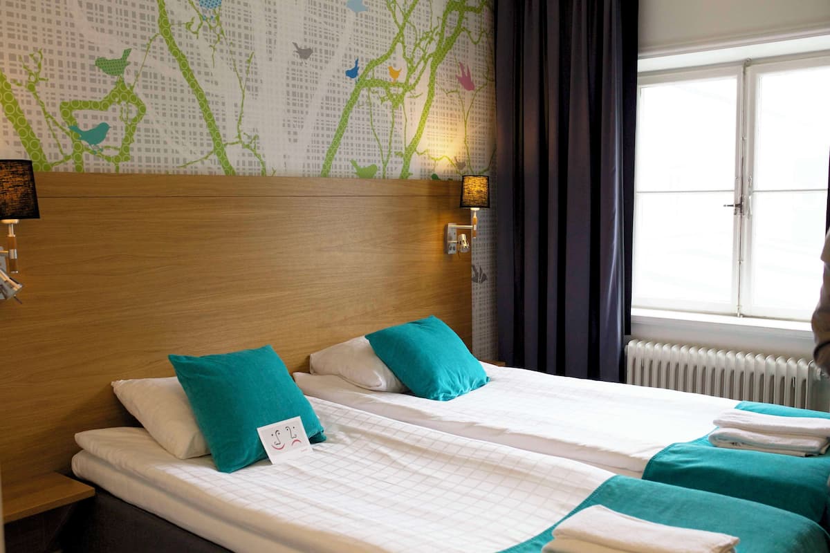 A hotel room with two beds, a wooden headboard, a wall with a nature-themed wallpaper, and a window with curtains.