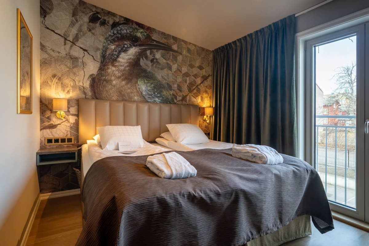 A hotel room with a large bed, two bedside tables, a stone wall mural, and a view of the outside.