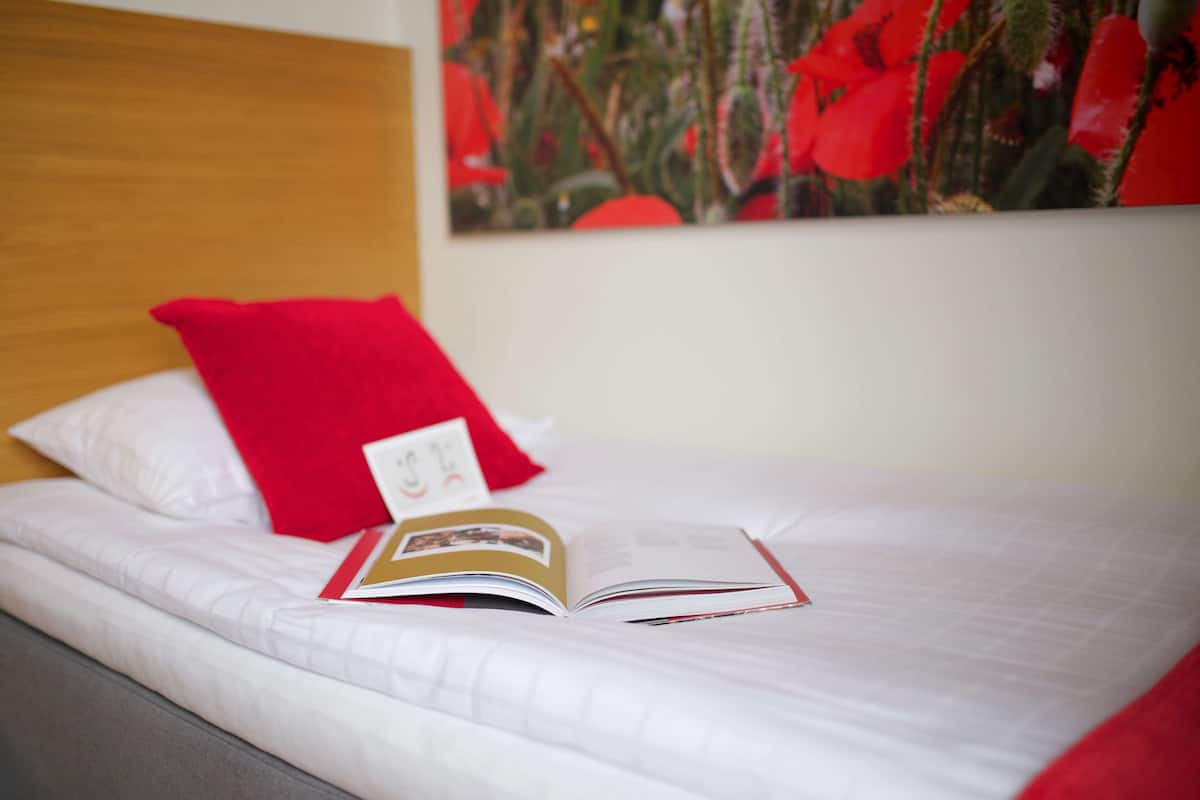 A neatly made bed with white bedding and a red pillow, an open book on the bed, and a red flower painting on the wall.