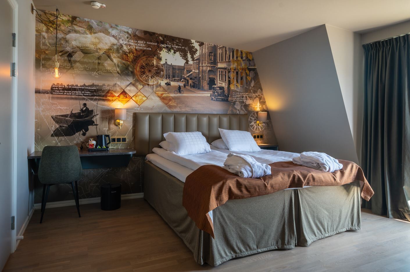 Una habitación de hotel moderna con una cama grande, un escritorio y una silla. En la pared, un mural que representa una escena histórica.
