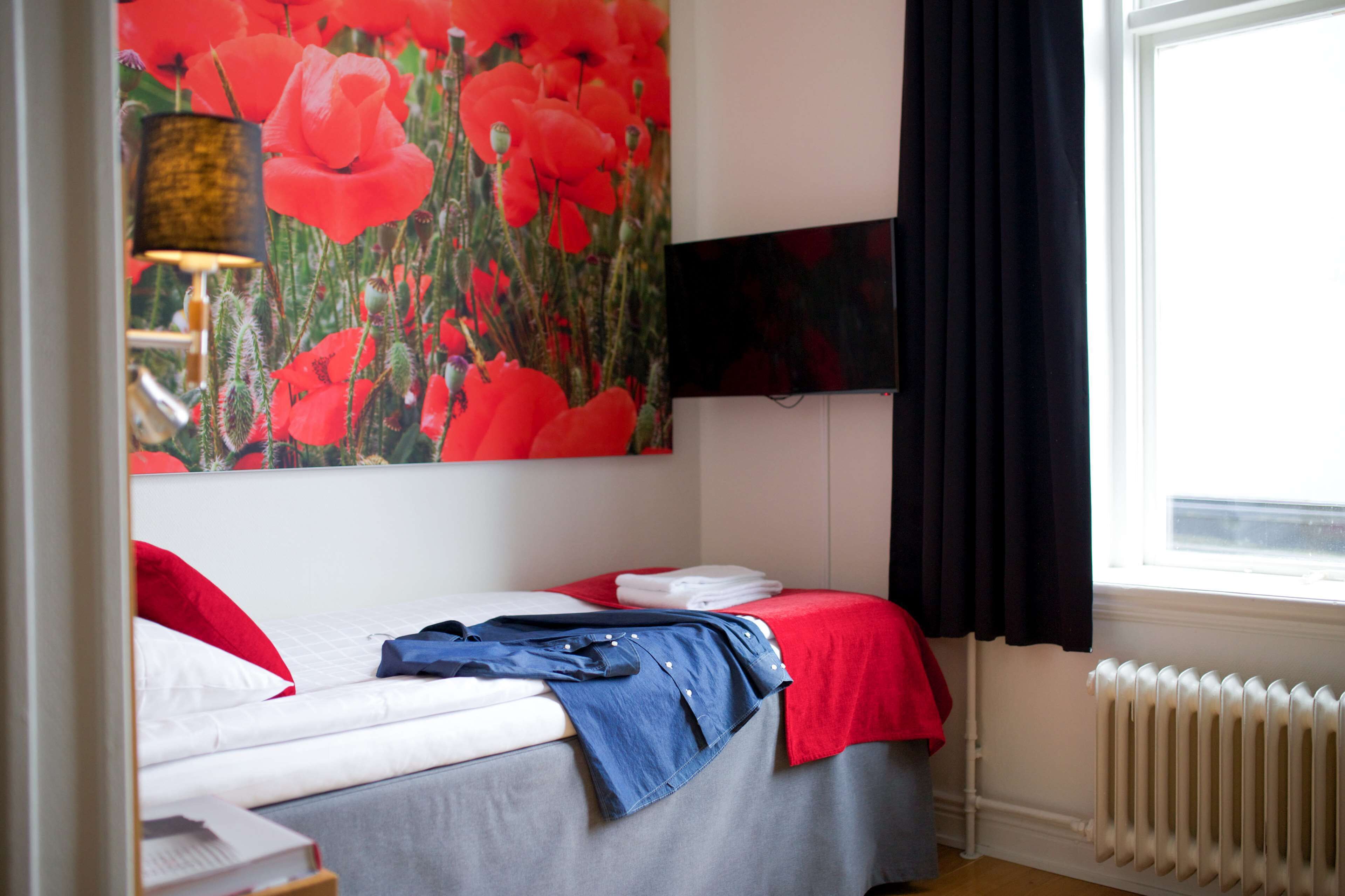 A hotel room with a bed, a red and white bedspread, a blue blanket, a red pillow, a black lamp, and a large red flower painting on the wall.
