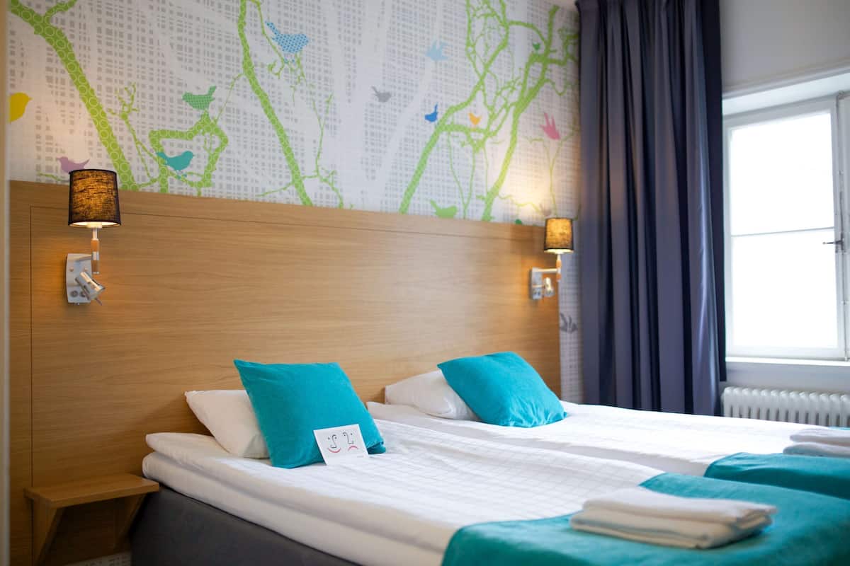 A hotel room with two beds, a wooden headboard, wall-mounted lamps, and a window with curtains.