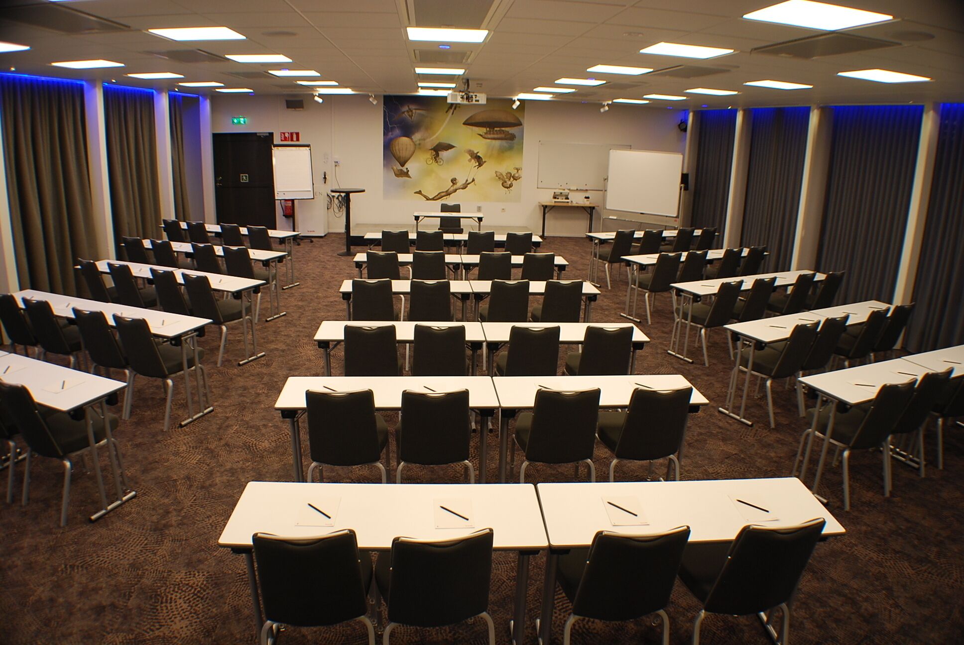 Une salle de conférence avec des rangées de tables et de chaises, un tableau blanc et un écran de projection.