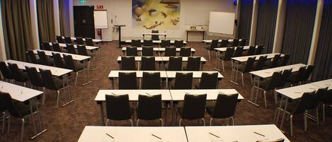 Une salle de conférence avec des rangées de tables et de chaises, un tableau blanc et un écran de projection.