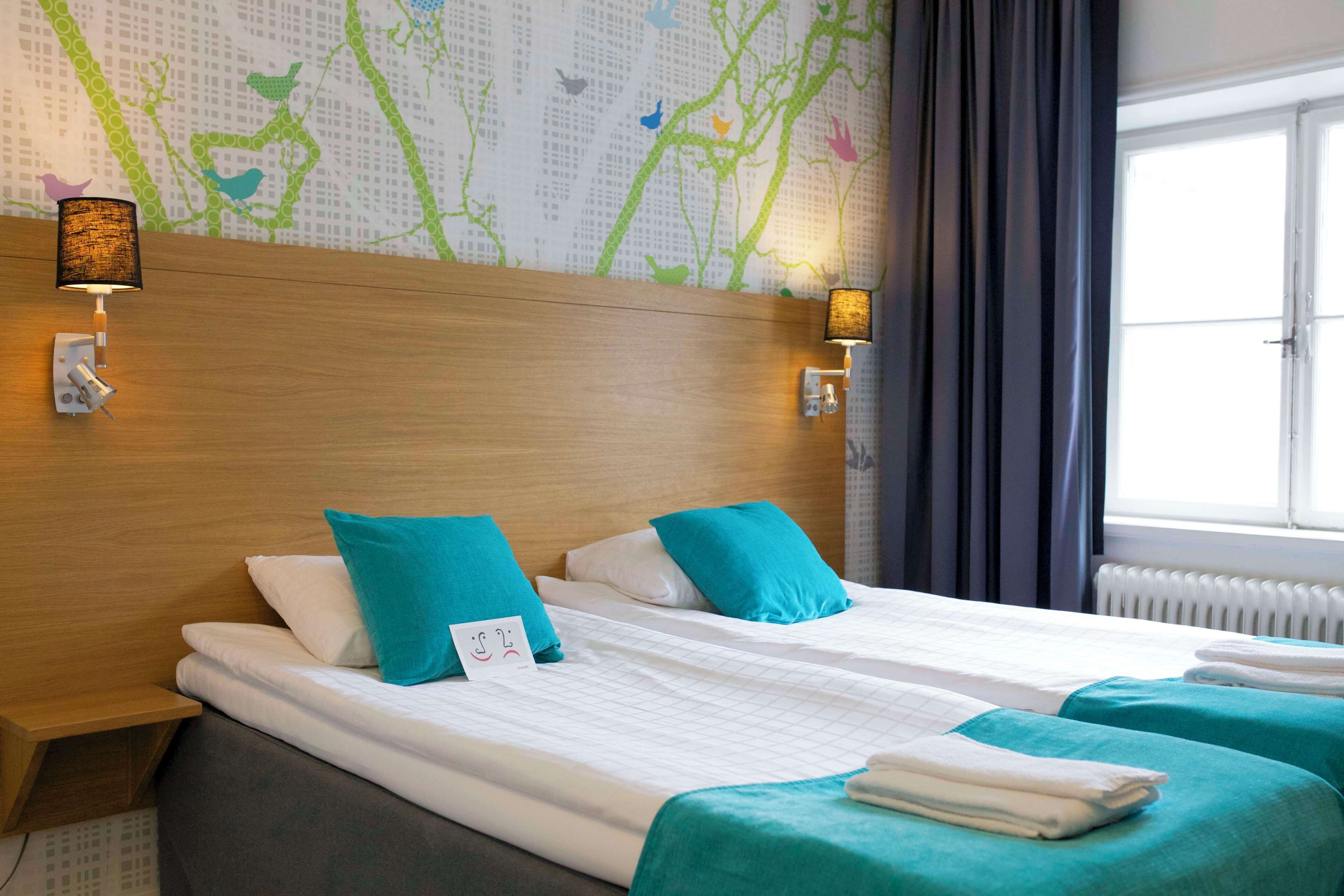 Habitación de hotel con cabecera de madera, dos camas con ropa de cama color turquesa, un mural en la pared y una ventana con cortinas.