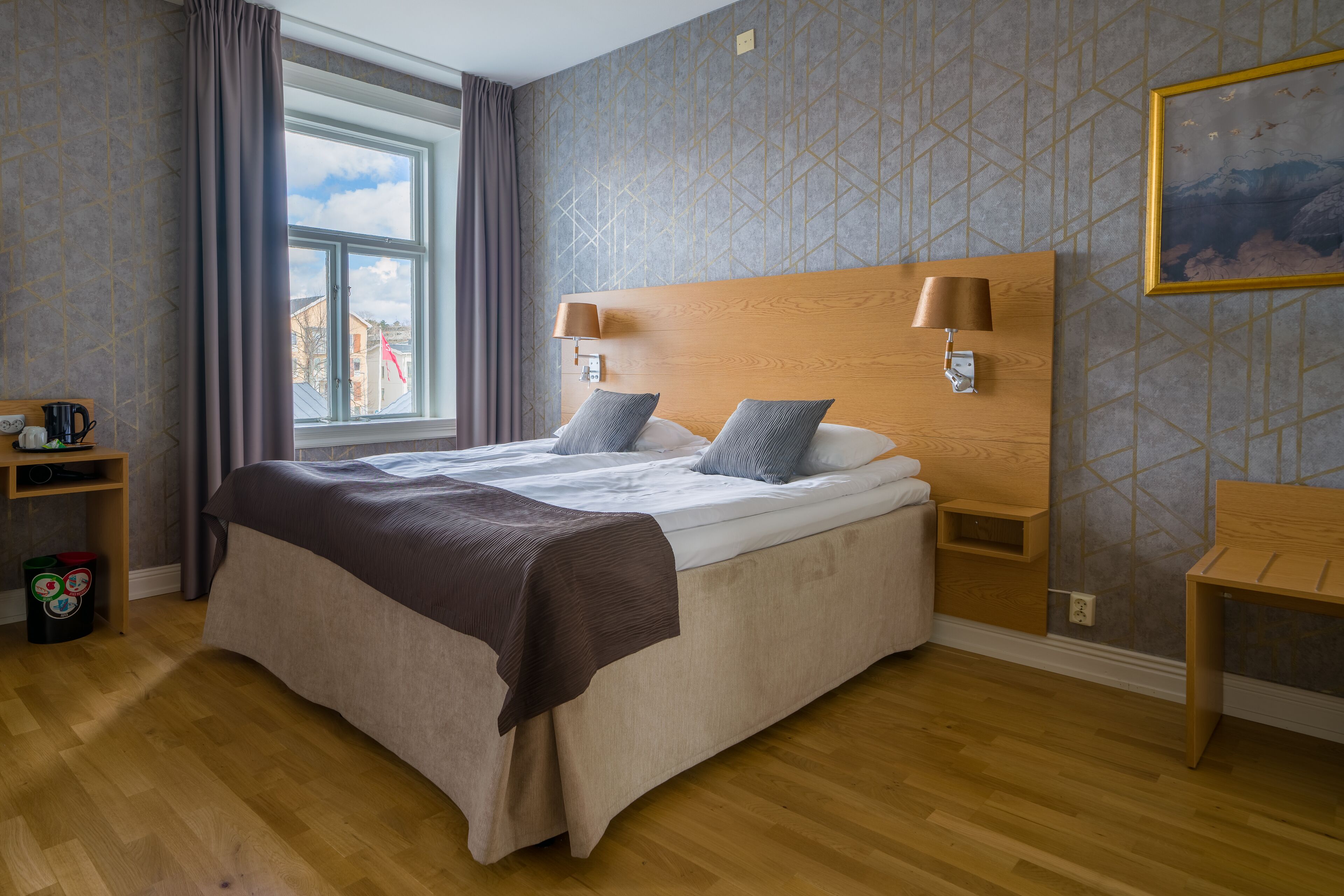 A neatly made bed with a wooden headboard, a bedside table with lamps, and a window with curtains.