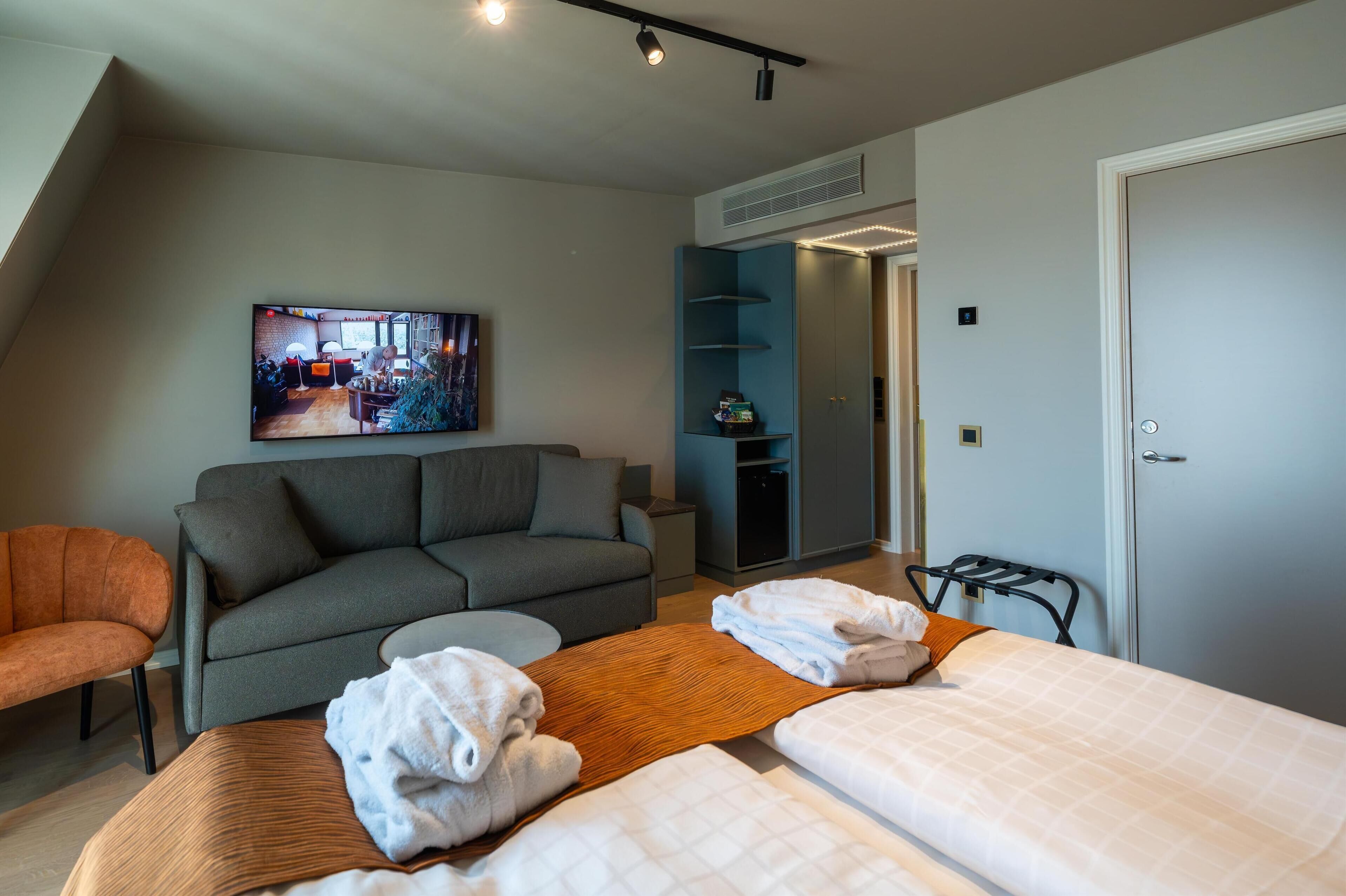 A modern hotel room with a gray sofa, a bed with white linens, a TV mounted on the wall, and a small table with towels.