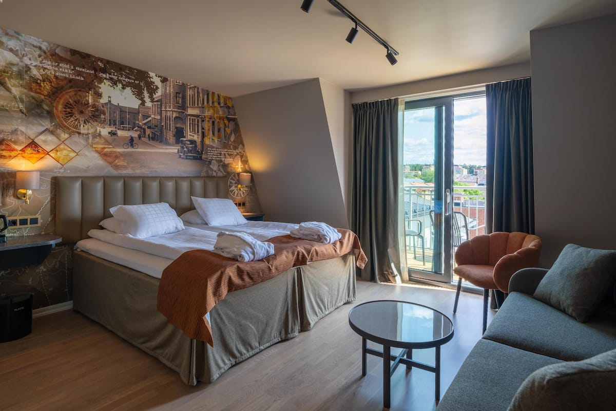 A hotel room with a bed, a sofa, a small table, and a view of the city through the window.