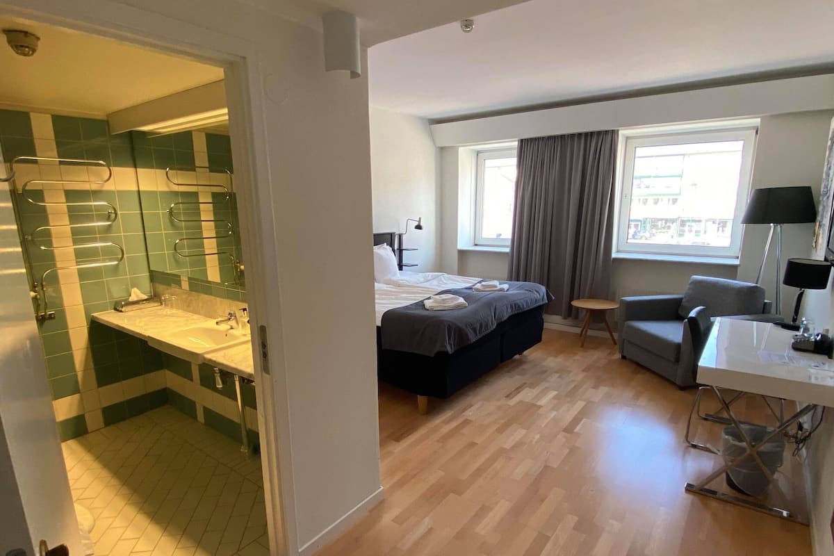 Une chambre d’hôtel comprenant un lit, un bureau, une chaise, une fenêtre et une salle de bain avec un miroir et un porte-serviettes.