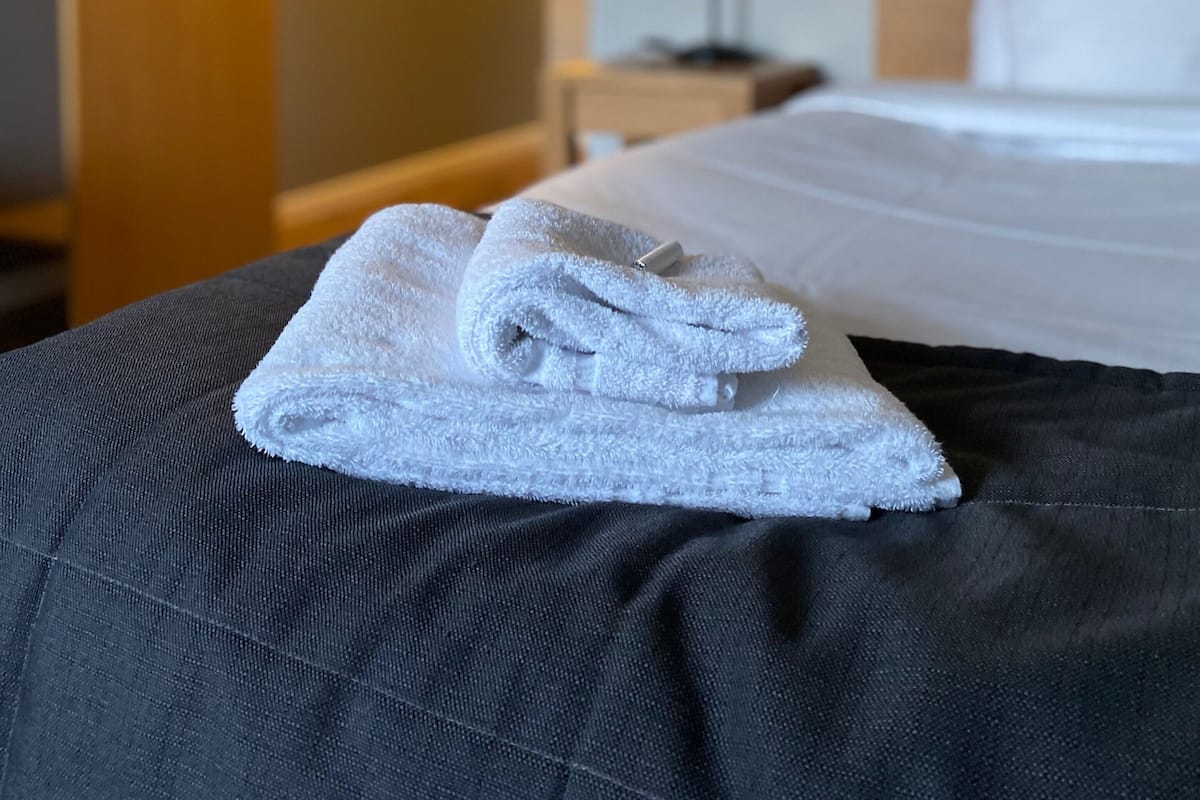 Un lit bien fait, avec une serviette blanche pliée posée sur la tête de lit, une lampe de chevet et une table de nuit avec une autre lampe.