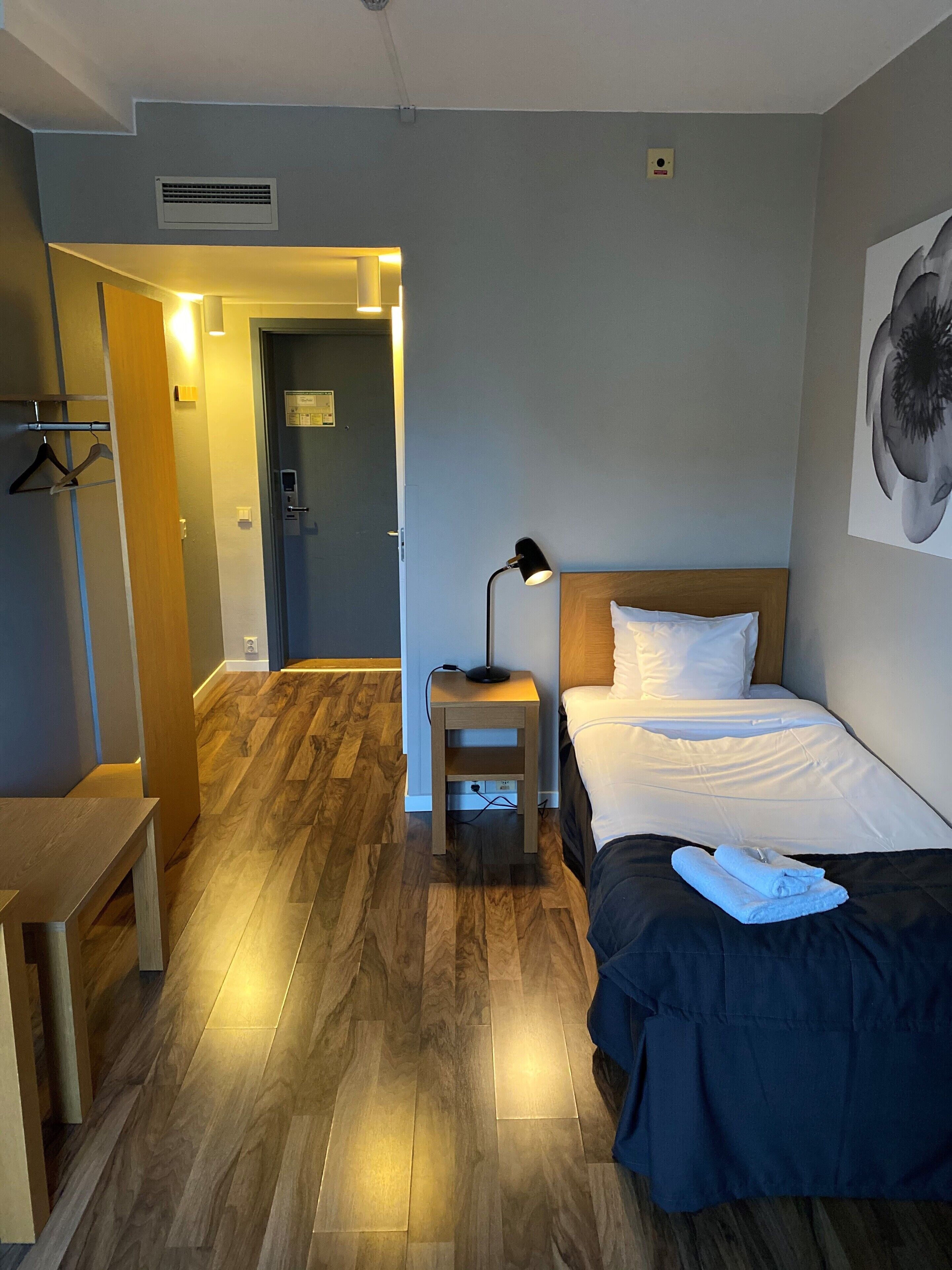 A hotel room with a wooden floor, a bed with white linens, a bedside table with a lamp, and a hanging coat rack.