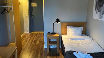 A hotel room with a wooden floor, a bed with white linens, a bedside table with a lamp, and a hanging coat rack.