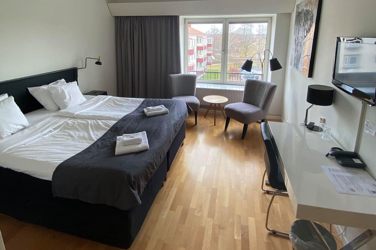 Une chambre d’hôtel avec un grand lit, deux fauteuils, une petite table ronde, un bureau avec un ordinateur et une fenêtre donnant sur des immeubles.