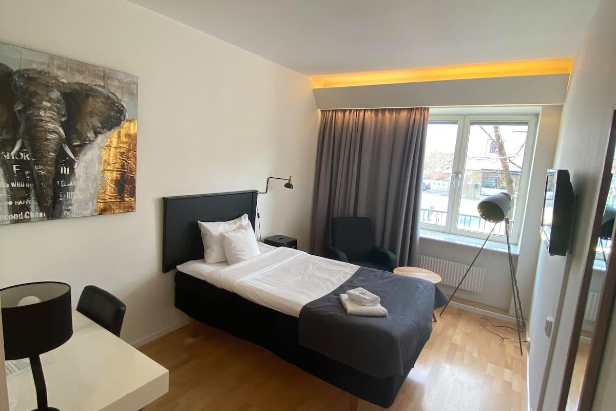 Une chambre d’hôtel avec un lit, un bureau, une chaise, une lampe, une fenêtre avec des rideaux et un tableau au mur.
