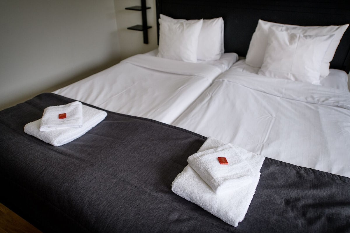 Une chambre d’hôtel avec deux lits, chacun agrémenté de draps et d’oreillers blancs, ainsi qu’un couvre-lit de couleur foncée.