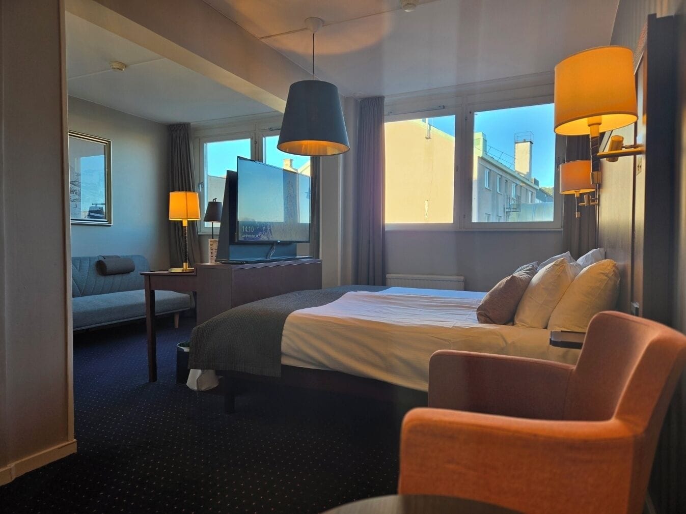 A hotel room with a bed, a desk, a television, and a window with a view of buildings.