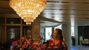 Une femme vêtue d’une robe à motifs floraux se tient dans un hall d’entrée, avec un grand lustre, un vase de fleurs et un comptoir d’accueil en arrière-plan.