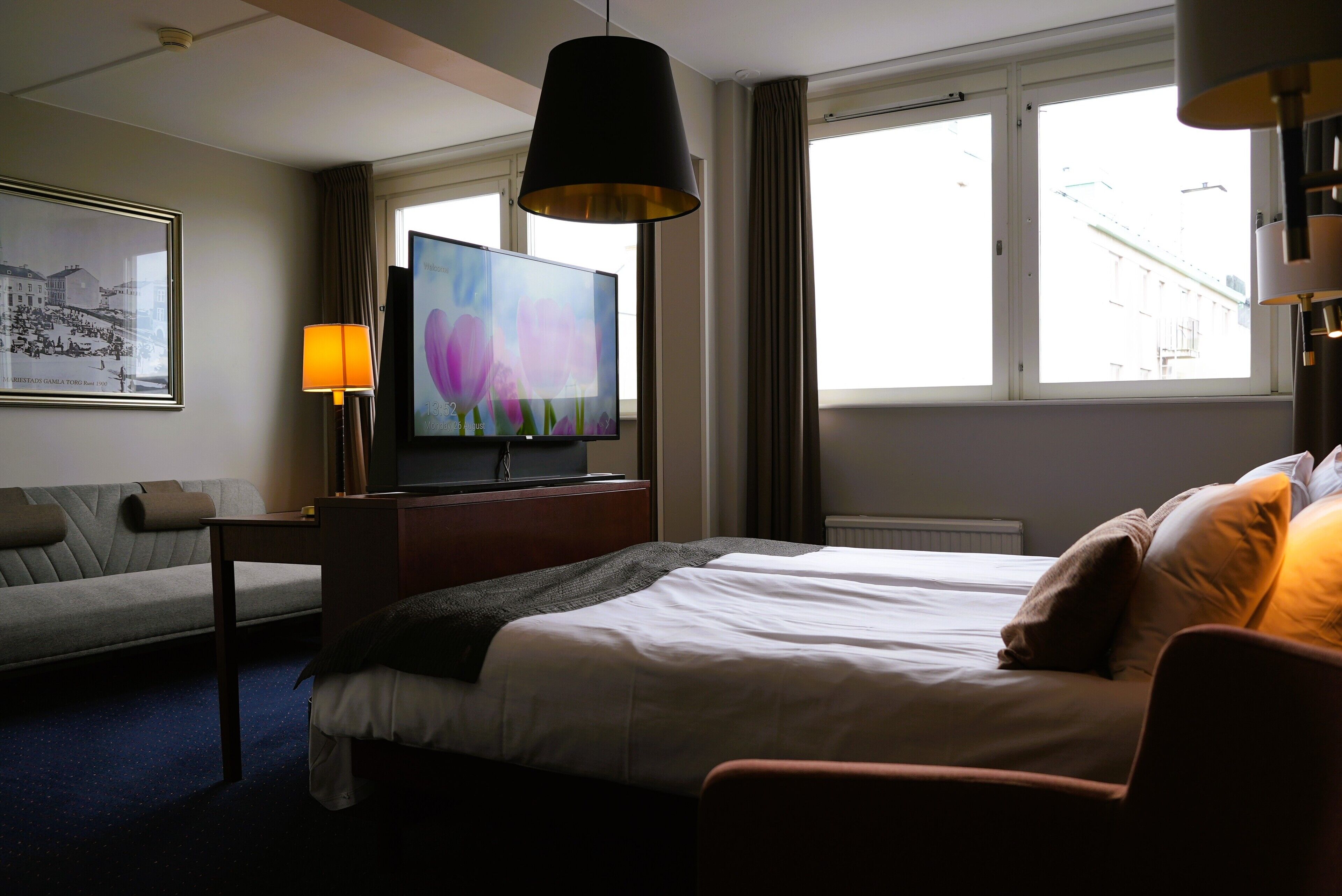 A hotel room with a bed, a TV, a sofa, a lamp, and a window with curtains.