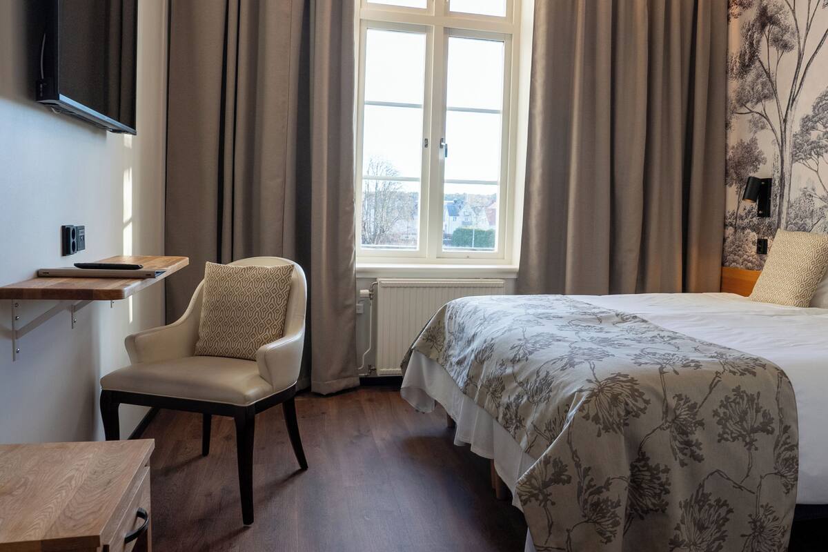 Un dormitorio con cama, una silla, una mesita, un ventanal y televisor instalado en la pared.