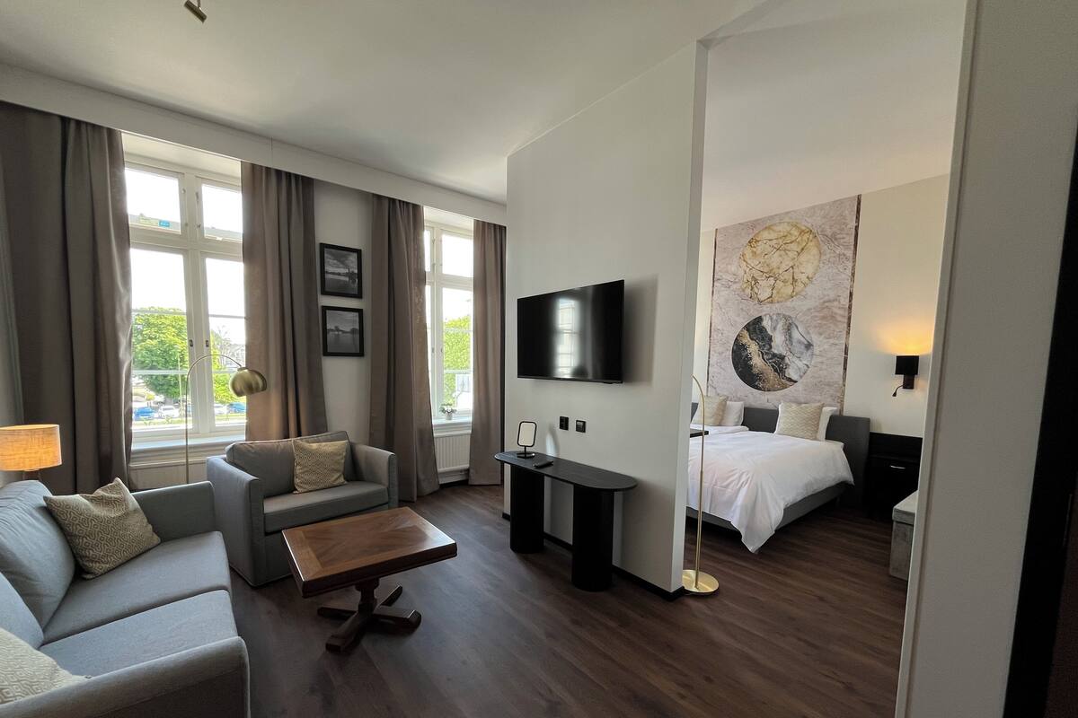 Habitación de hotel con sofá, mesa de centro, televisión, cama y un amplio ventanal con cortinas.