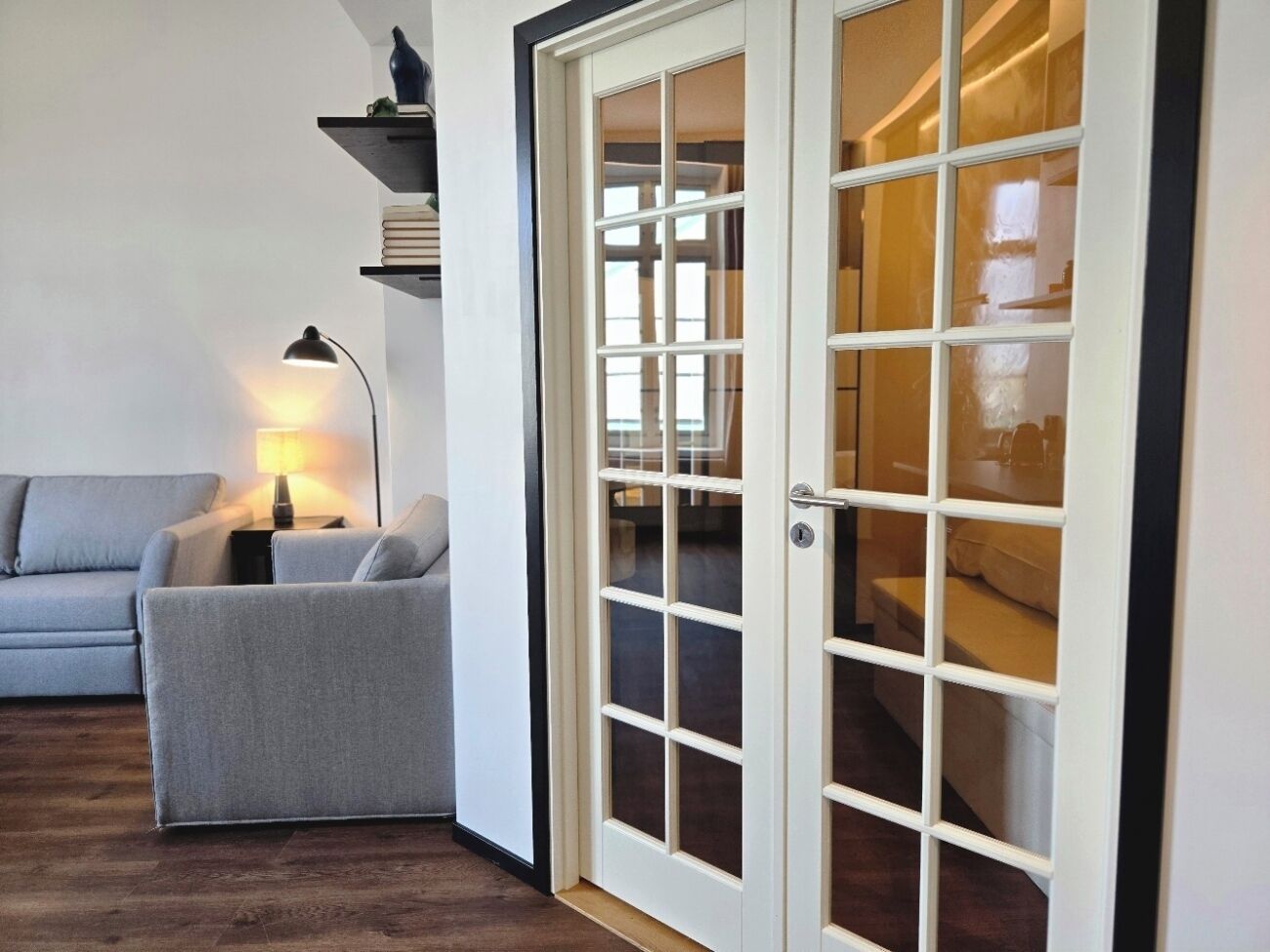 Una habitación con un sofá gris, una lámpara y una puerta blanca con paneles de vidrio.