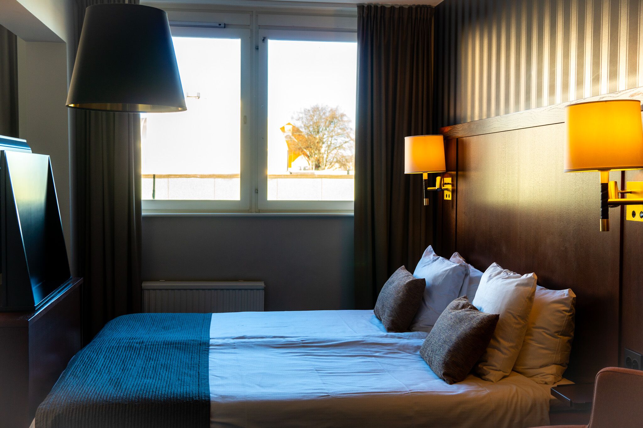 Une chambre d’hôtel équipée d’un lit, d’une télévision, d’une fen être avec des rideaux et de deux lampes fixées au mur.