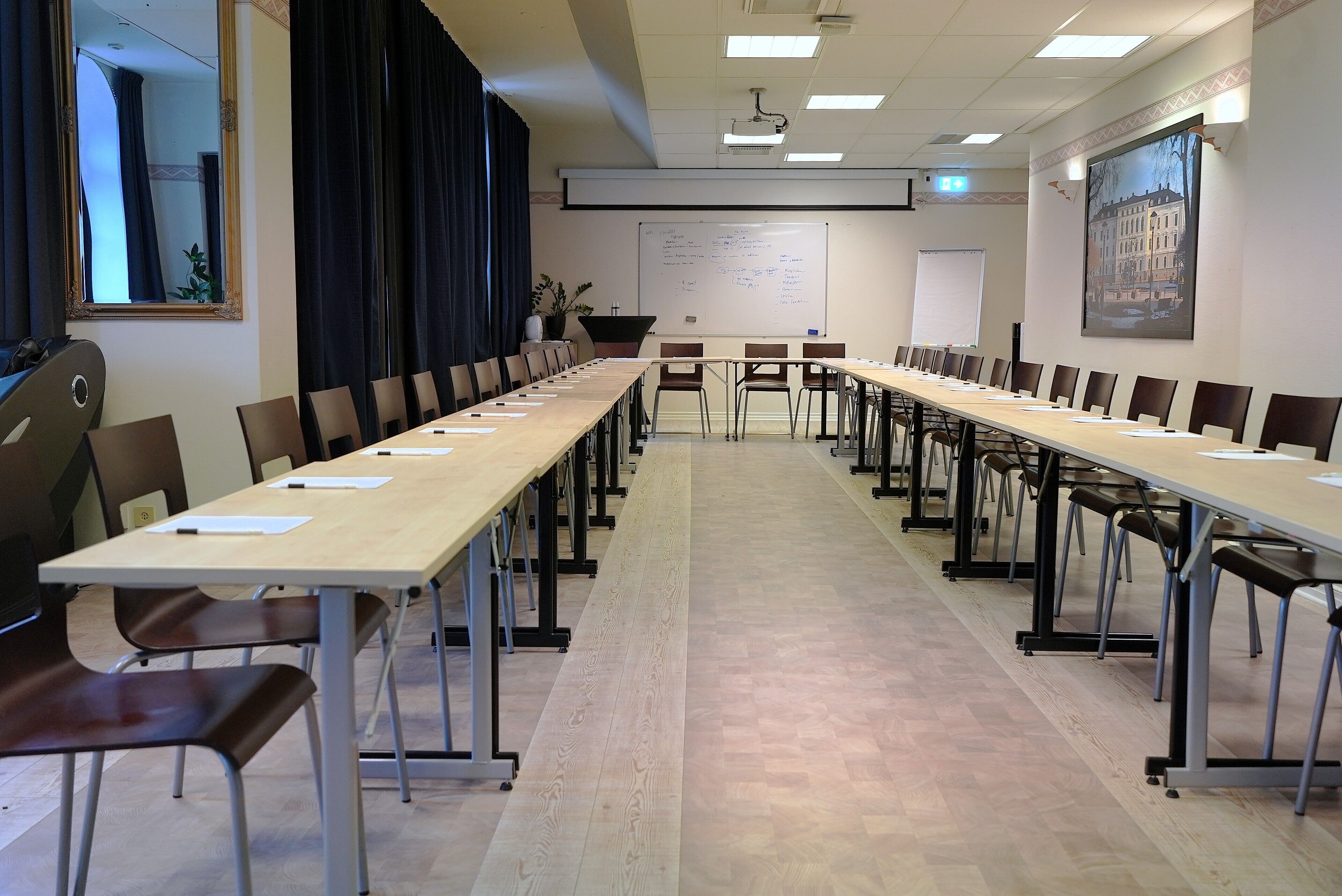 Un salón de conferencias con largas mesas, sillas, una pizarra y una pantalla para proyector.