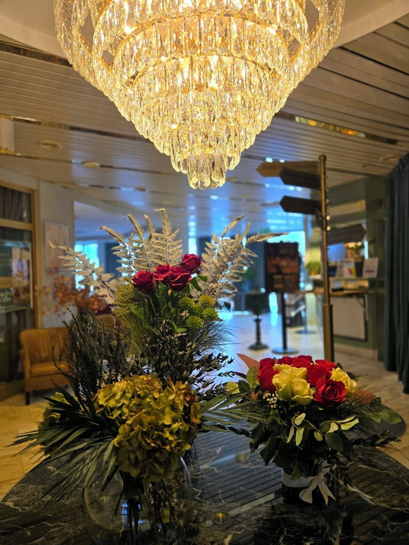A lobby with a large chandelier, two floral arrangements on a marble table, and a modern interior design.