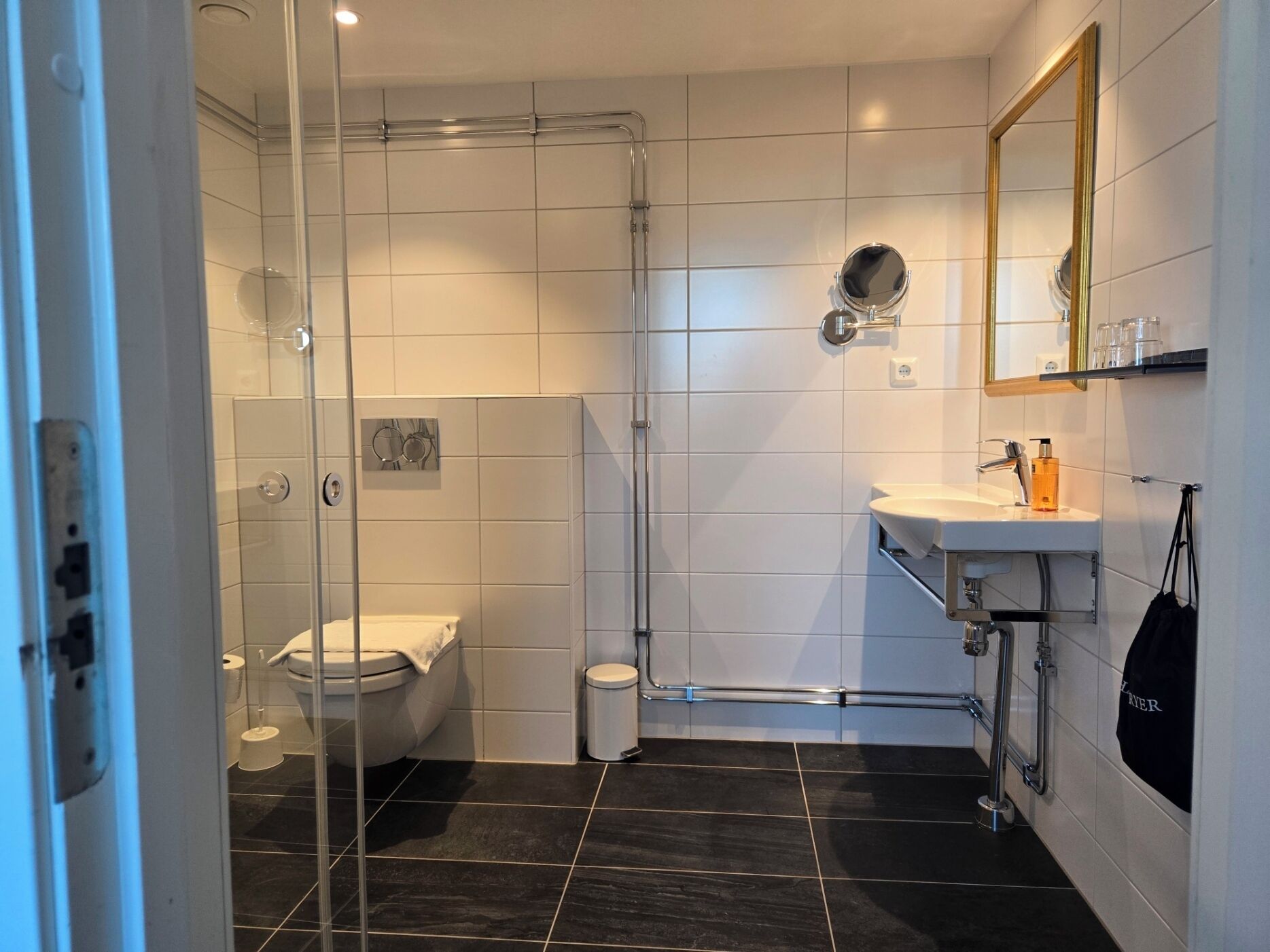 A modern bathroom with a glass-enclosed shower, a white toilet, a sink with a mirror, and a black-tiled floor.