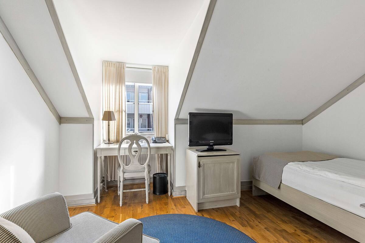 Una habitación pequeña y bien iluminada, con escritorio, silla, televisor y cama.