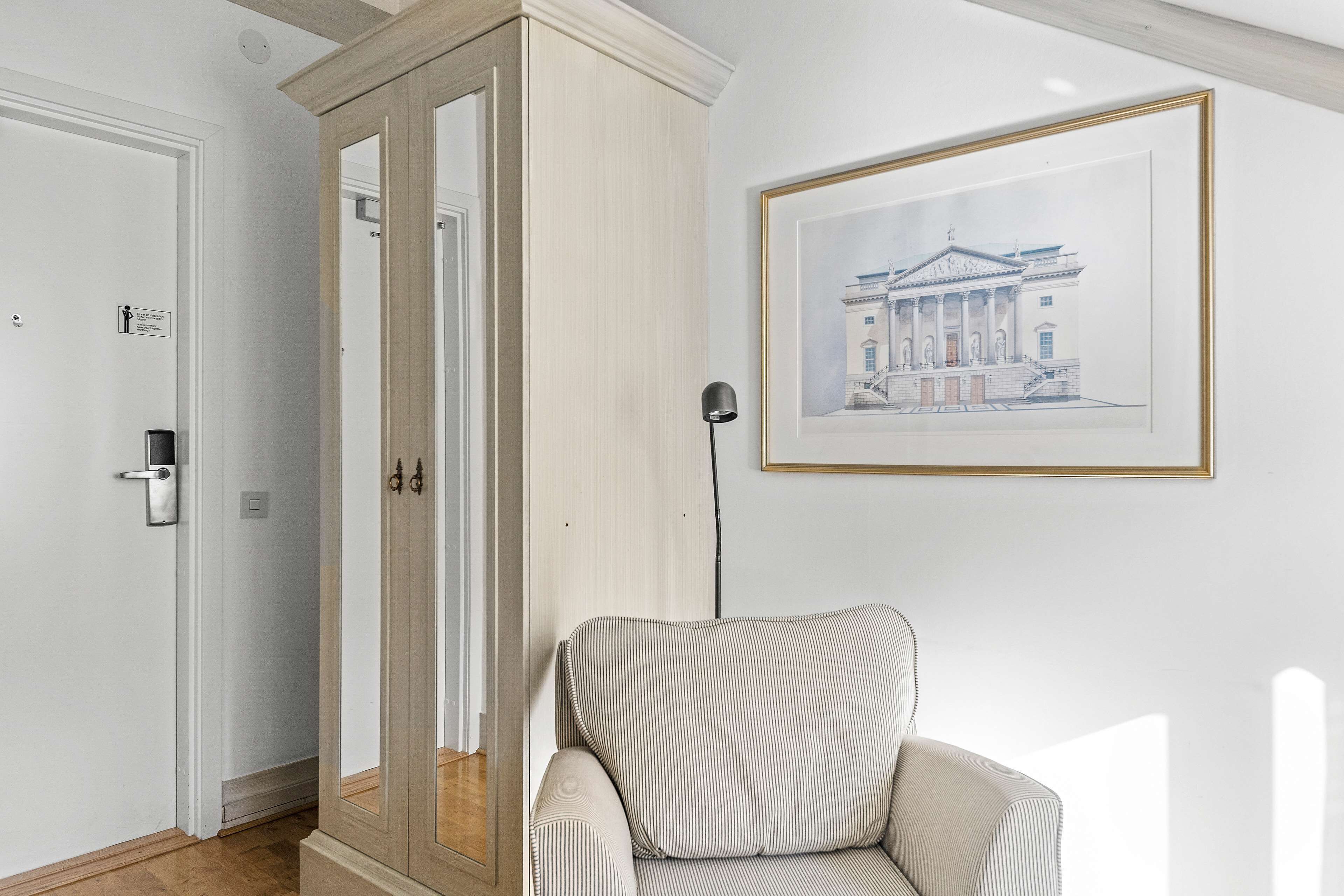 A room with a striped armchair, a framed architectural drawing on the wall, and a wooden cabinet with glass doors.