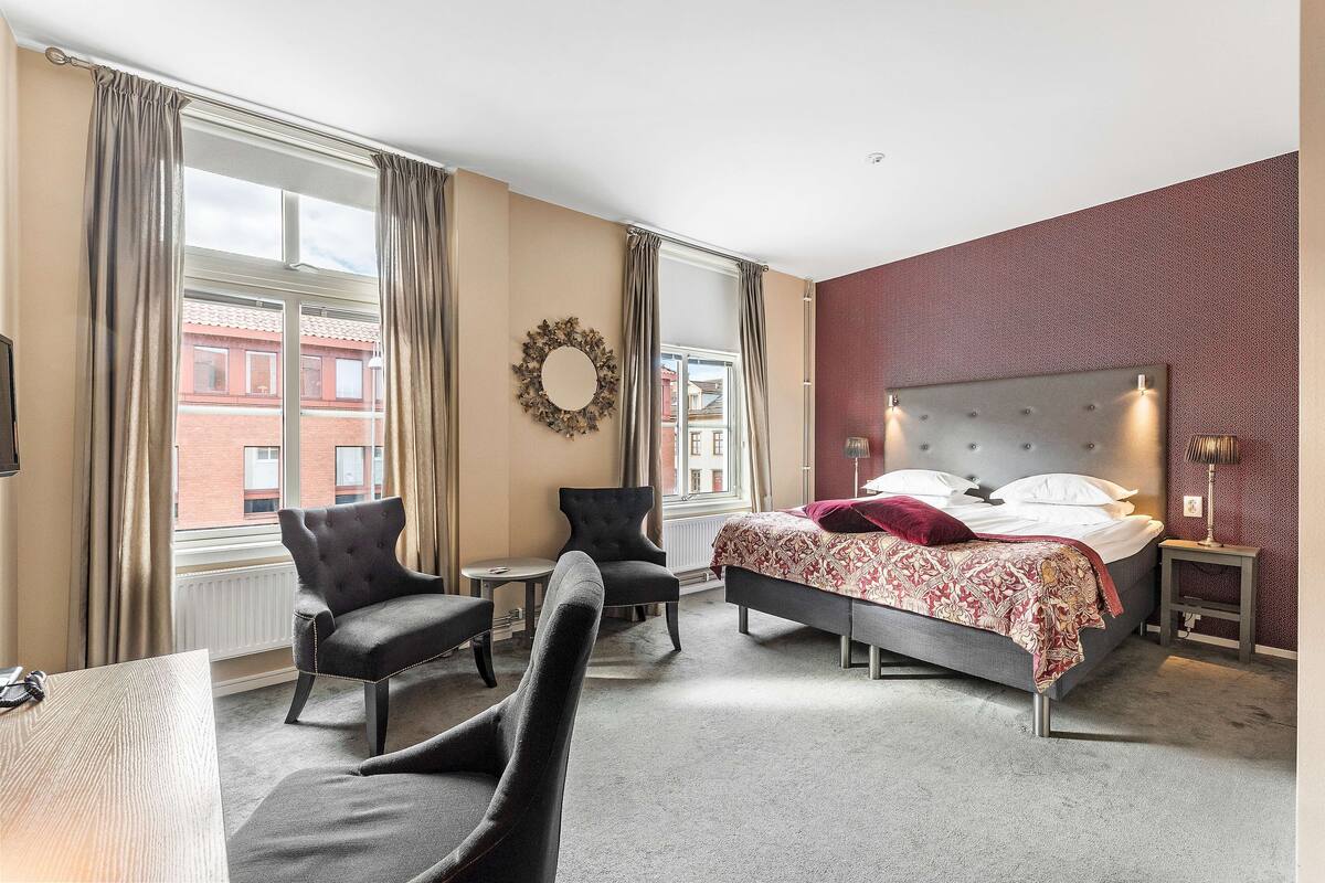 Habitación de hotel con cama, dos sillones, una mesita y vistas a un edificio de ladrillo rojo a través del ventanal.