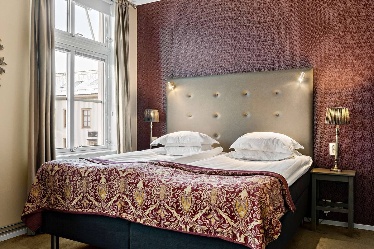 Una cama doble con colcha estampada, cabecera capitonada, lámparas de noche y una ventana con cortinas.
