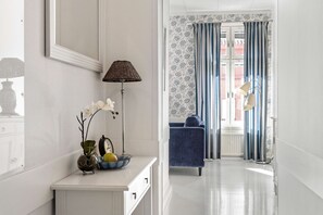 Un pasillo con una consola blanca, una lámpara y un jarrón con flores. Se ve una silla azul y una puerta con cortinas estampadas.