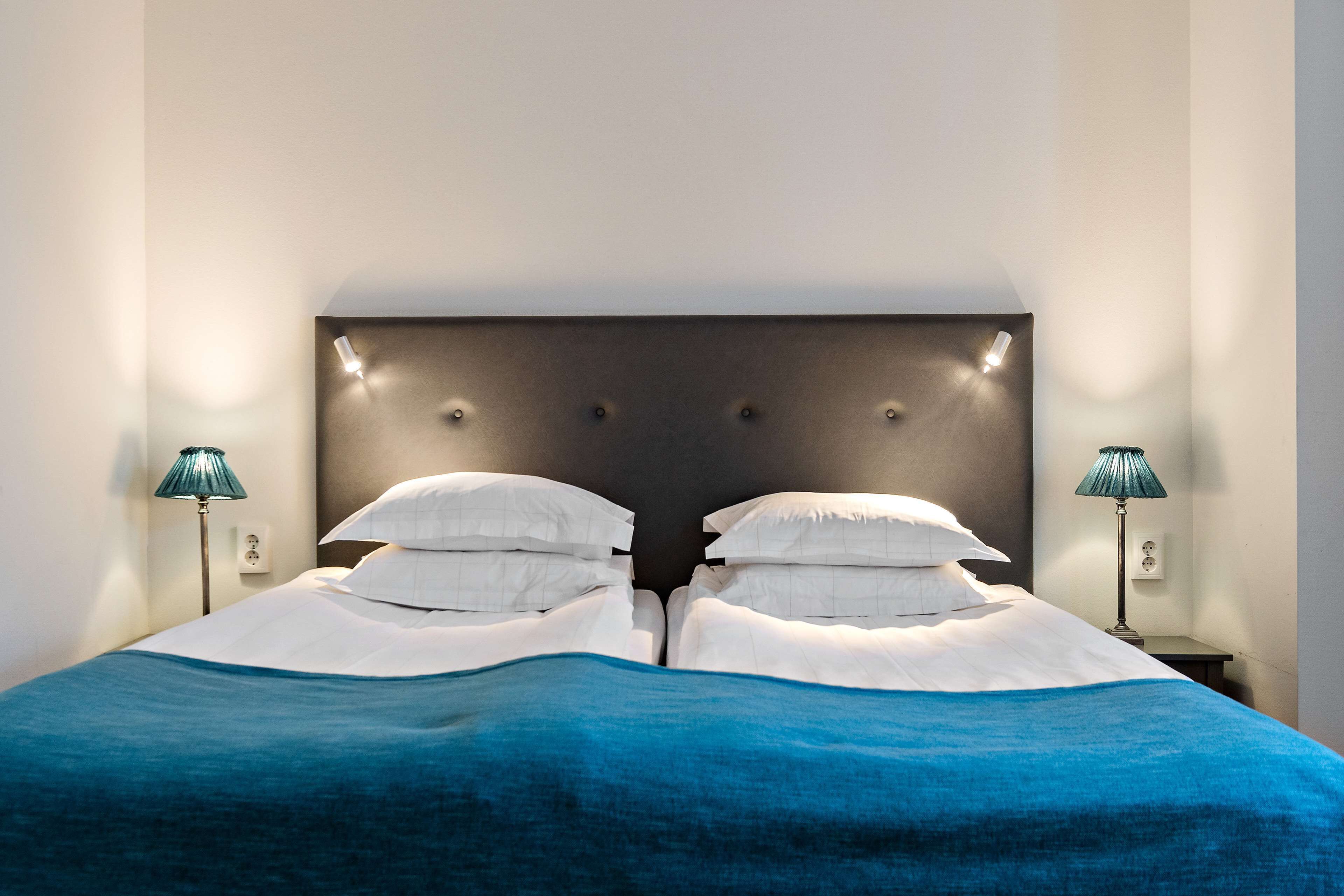 Een net opgemaakt bed met een blauwe sprei en witte kussens, met aan beide kanten nachtlampjes.