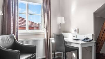 A small, modern room with a white desk, a black leather chair, a lamp, and a view of a red-tiled roof outside.