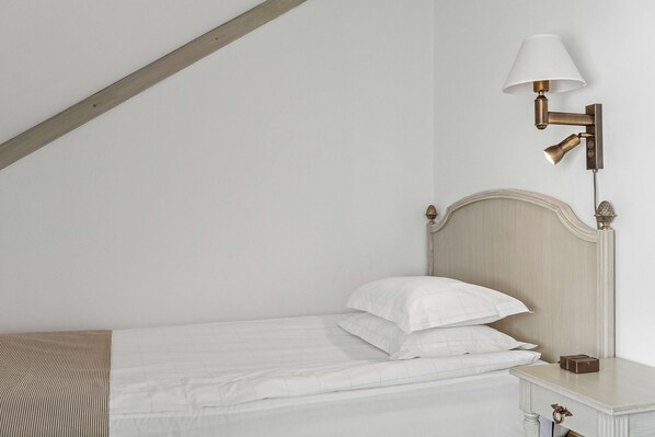 Una cama bien hecha con ropa de cama y almohadas blancas, una mesita de noche con una lámpara, y una viga de techo de madera.