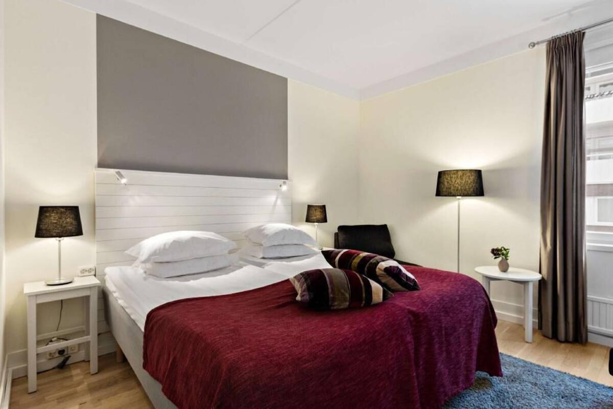 Una habitación de hotel con una cama, dos mesitas de noche, una lámpara, una silla, una pequeña mesa con una planta y una ventana con cortinas.