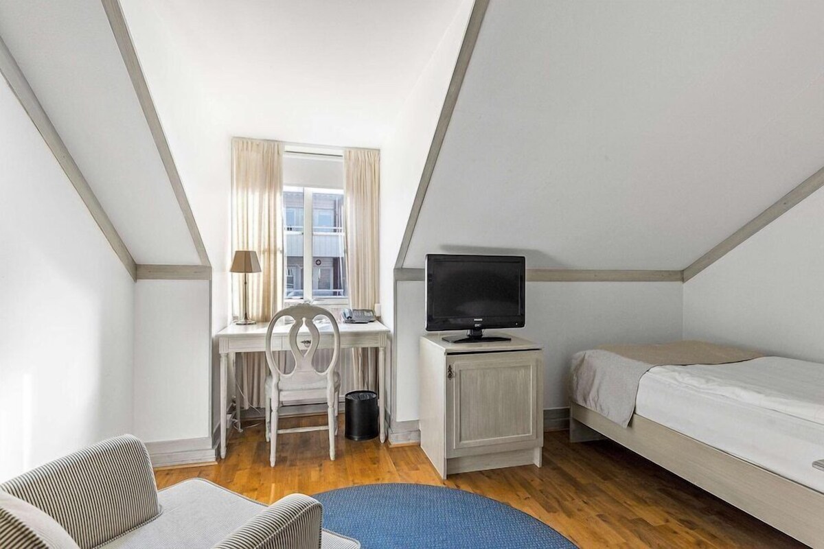 A small, well-lit room with a desk, chair, TV, and a bed.