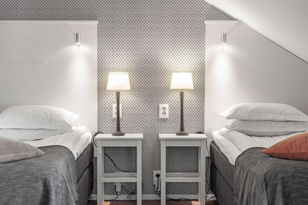 Un dormitorio con dos camas, cada una con una mesita de noche y una lámpara. La habitación tiene un papel tapiz estampado y un techo inclinado.