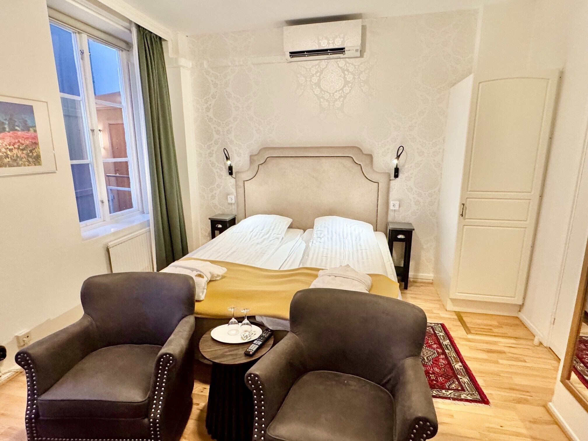 A hotel room with a bed, two armchairs, a small table with a remote control, and a window with curtains.