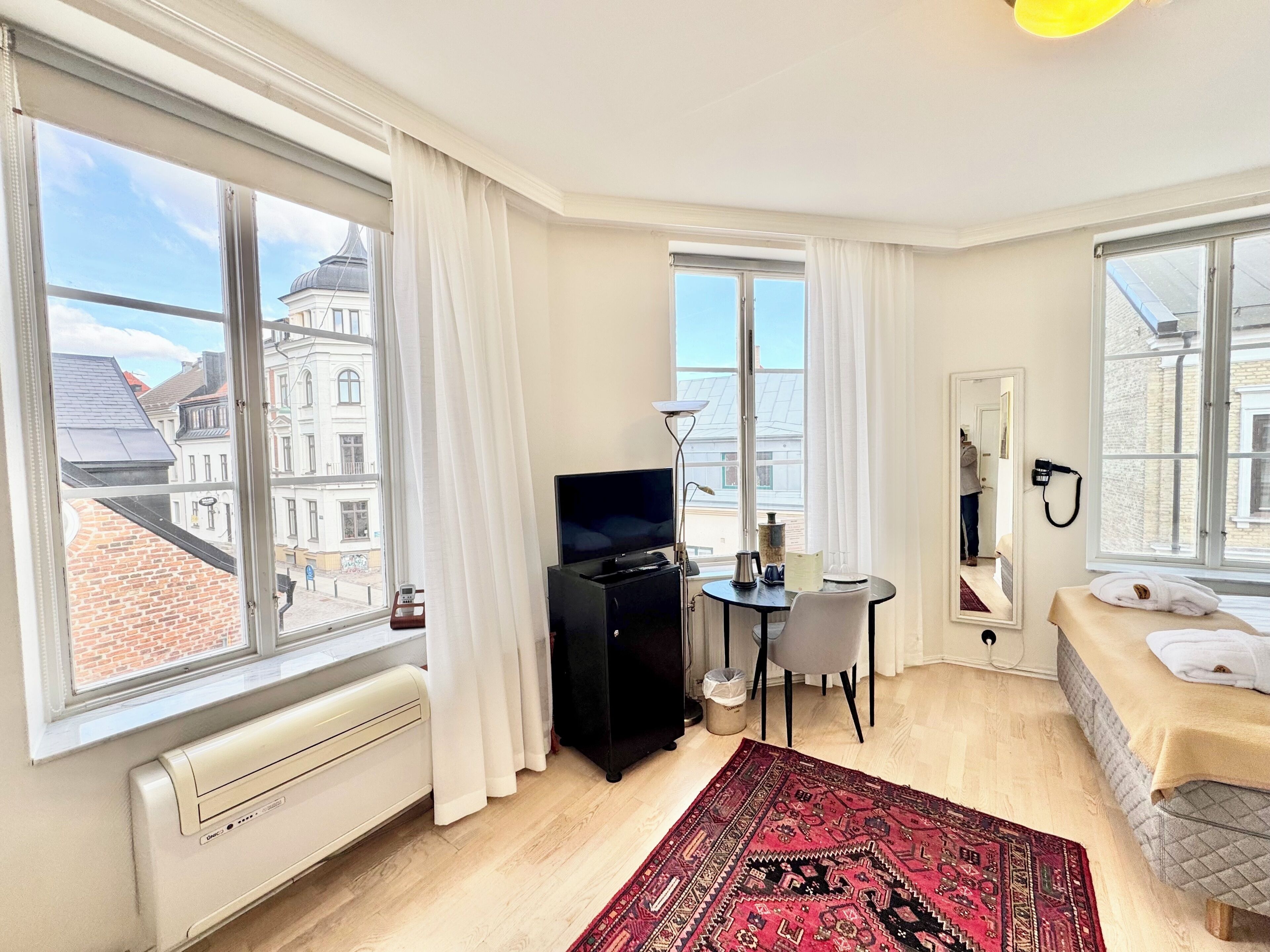 Habitació d'hotel amb grans finestres, un llit, escriptori amb cadira, televisió i radiador.