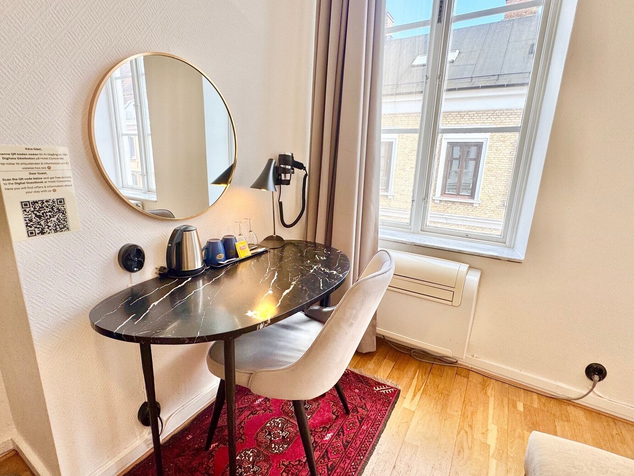A compact hotel room with a small round table, a chair, a mirror, a kettle, a telephone, and a window with a view of buildings.