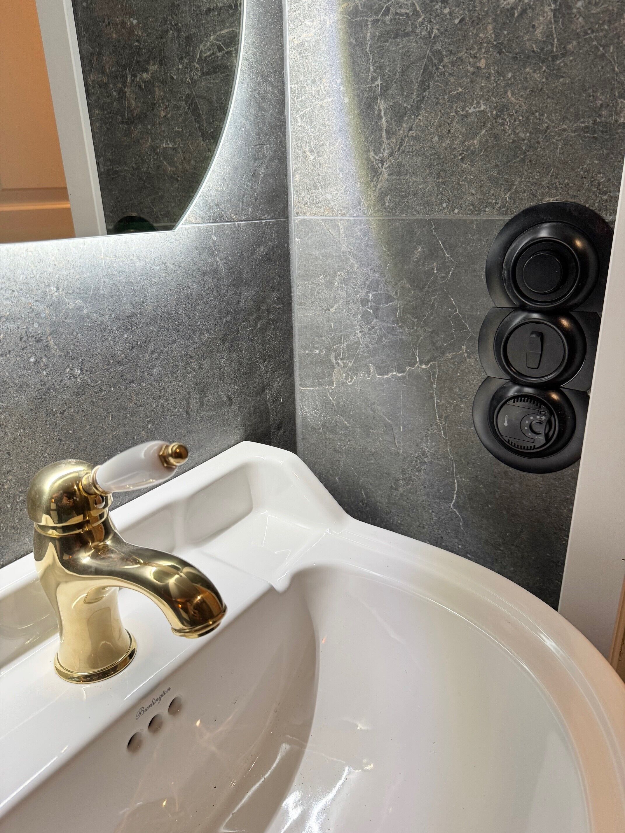 Un baño con un lavamanos blanco, grifo dorado y controles negros instalados en la pared.