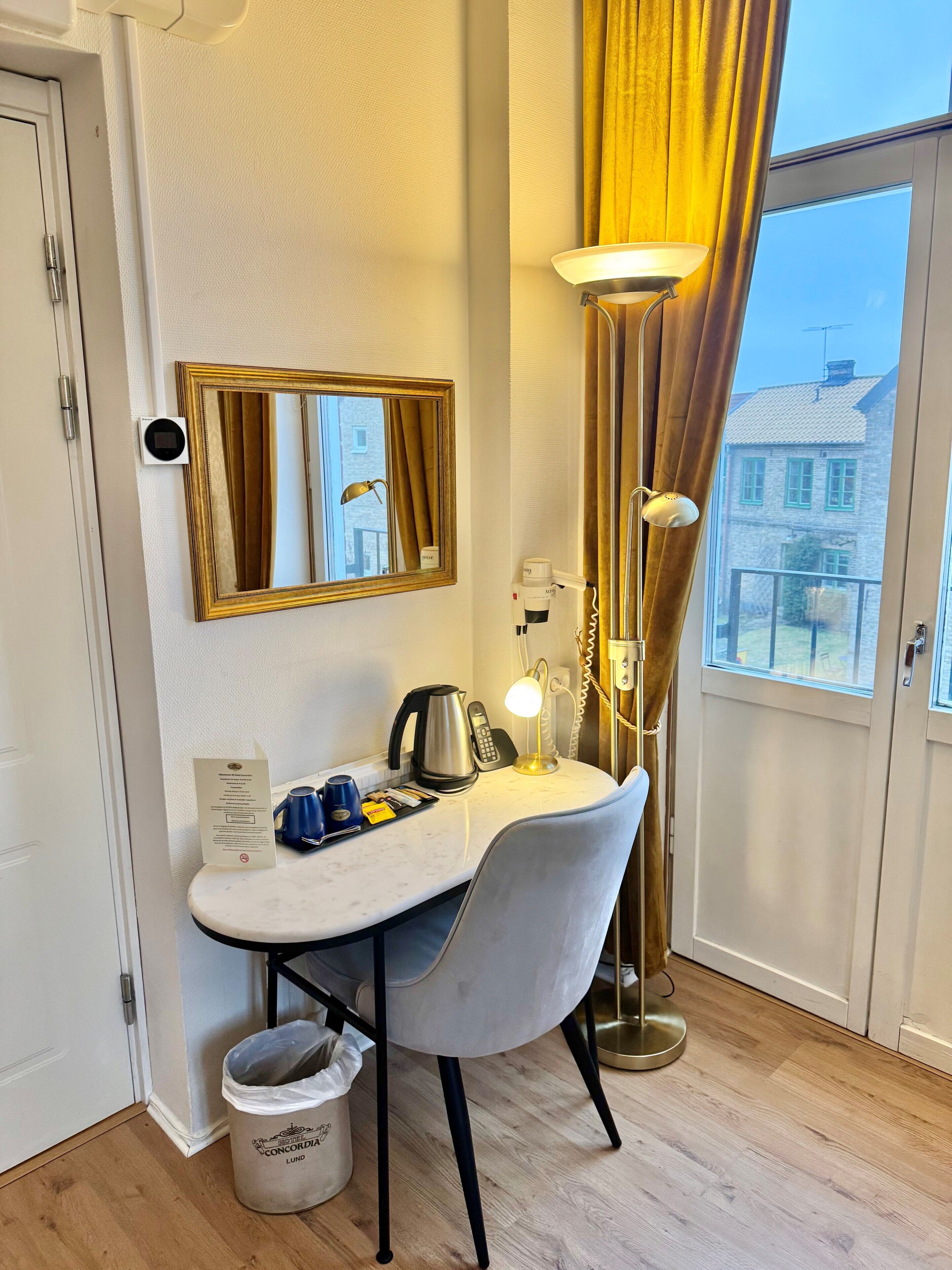 Habitación de hotel con tocador, espejo, lámpara, tetera, teléfono y vista a un edificio.