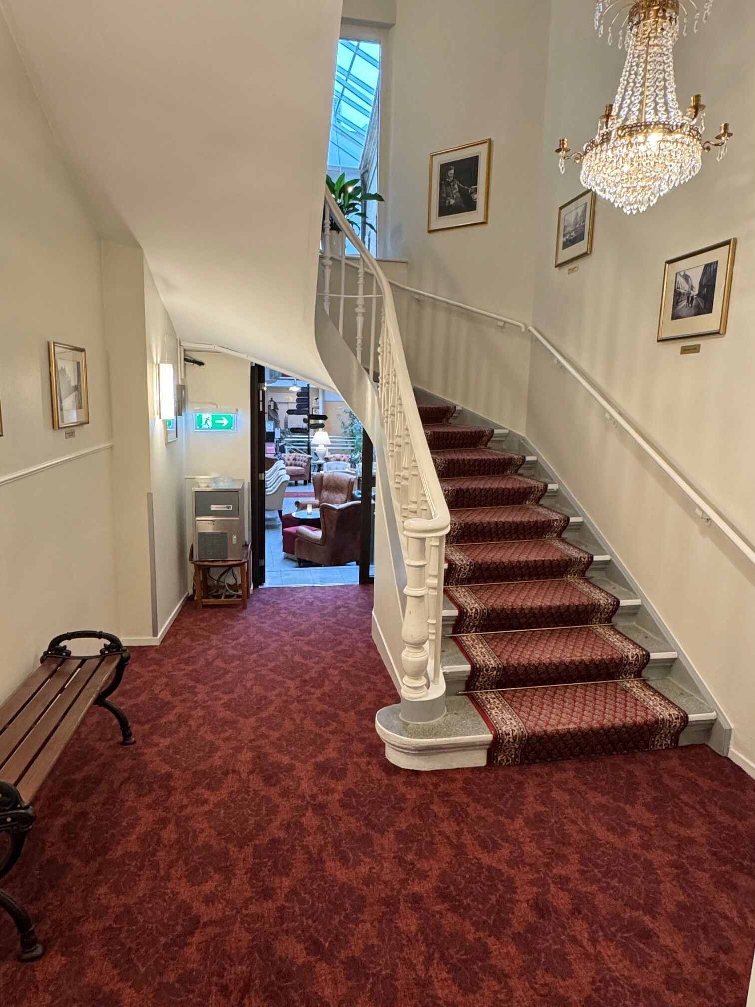 A grand staircase with a red carpet, a chandelier, framed pictures, and a bench.