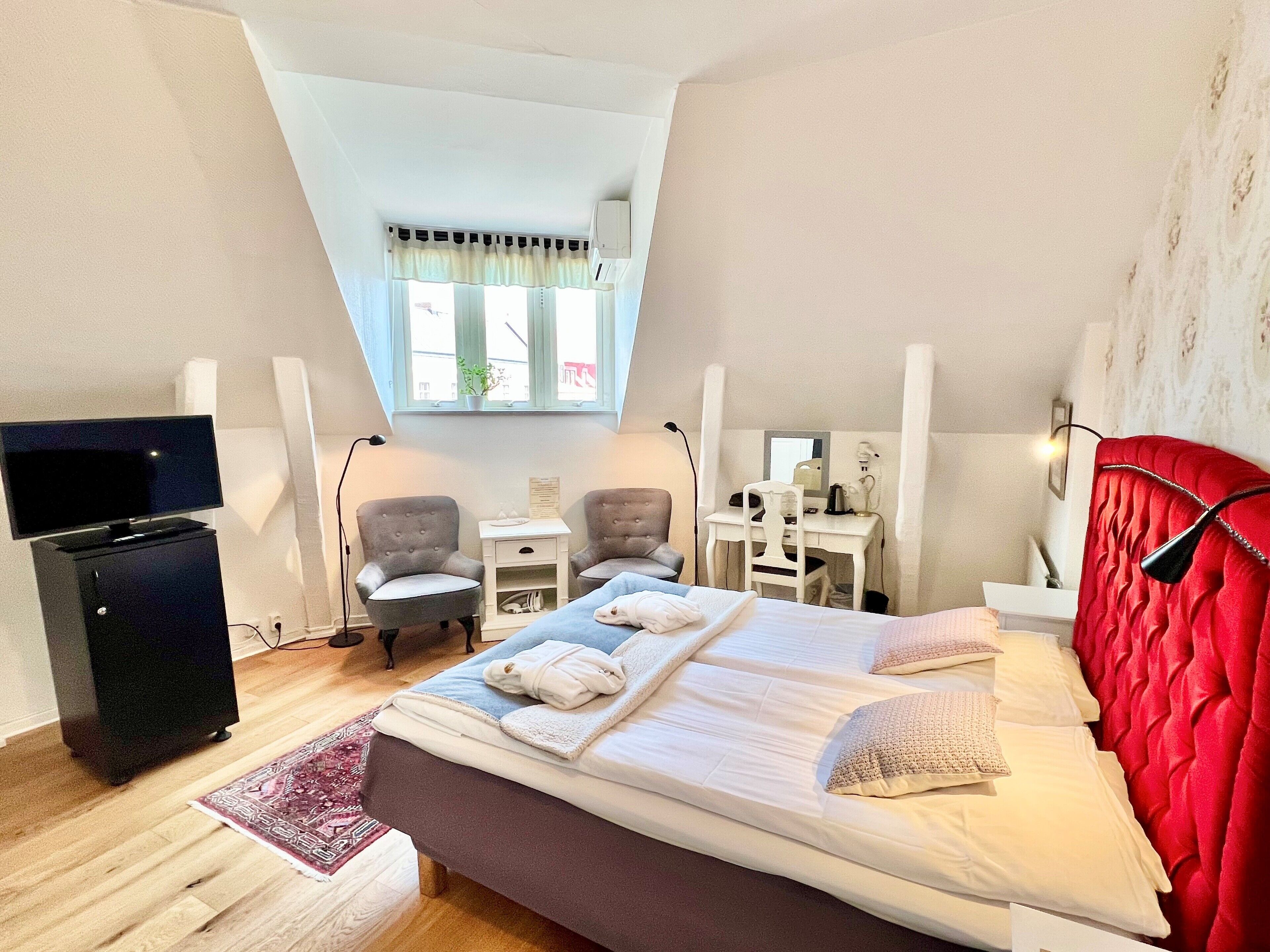 Une chambre d’hôtel avec un grand lit, deux fauteuils, un bureau avec un ordinateur, une télévision et un petit meuble de rangement.